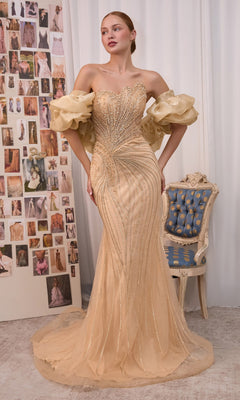 A woman wears the Andrea & Leo A1519 long gold prom dress, featuring a strapless sweetheart neckline, elegant beaded detailing, and dramatic puff sleeves, as she stands beside a vintage chair and a collage wall of fashion photos.