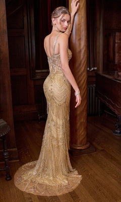 Wearing the Andrea & Leo A1526 Long Prom Dress, a woman in an ornate, floor-length gold gown adorned with beaded fringe stands with her back turned, one arm resting on a wooden column in an elegant, dimly lit room with wood paneling.