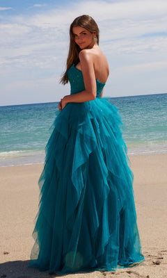 A woman in the Nox Anabel A1591 long prom dress—a strapless corset gown with a ruffled A-line skirt—stands on a sandy beach, facing the camera with the ocean and sky behind her.