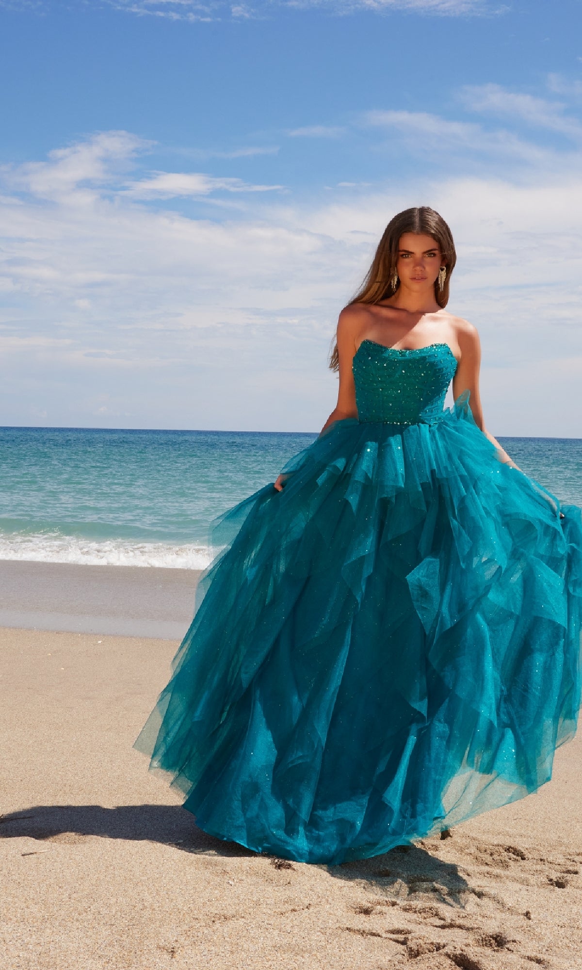 Wearing the Nox Anabel A1591 Long Prom Dress, a woman stands on a sandy beach, the turquoise strapless corset gown with a ruffled A-line skirt flowing beautifully against the ocean and blue sky.