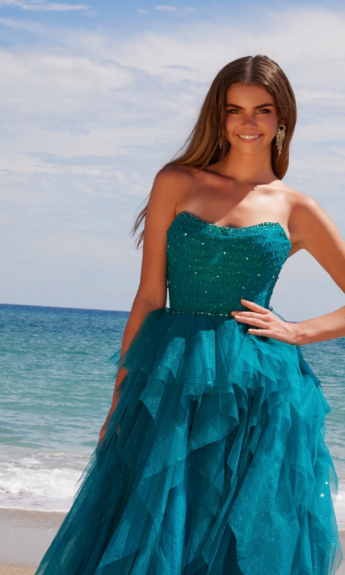 A woman in the Nox Anabel A1591 long prom dress, featuring a strapless corset bodice and ruffled teal tulle A-line skirt, stands on the beach with the shimmering ocean and sky as her backdrop.