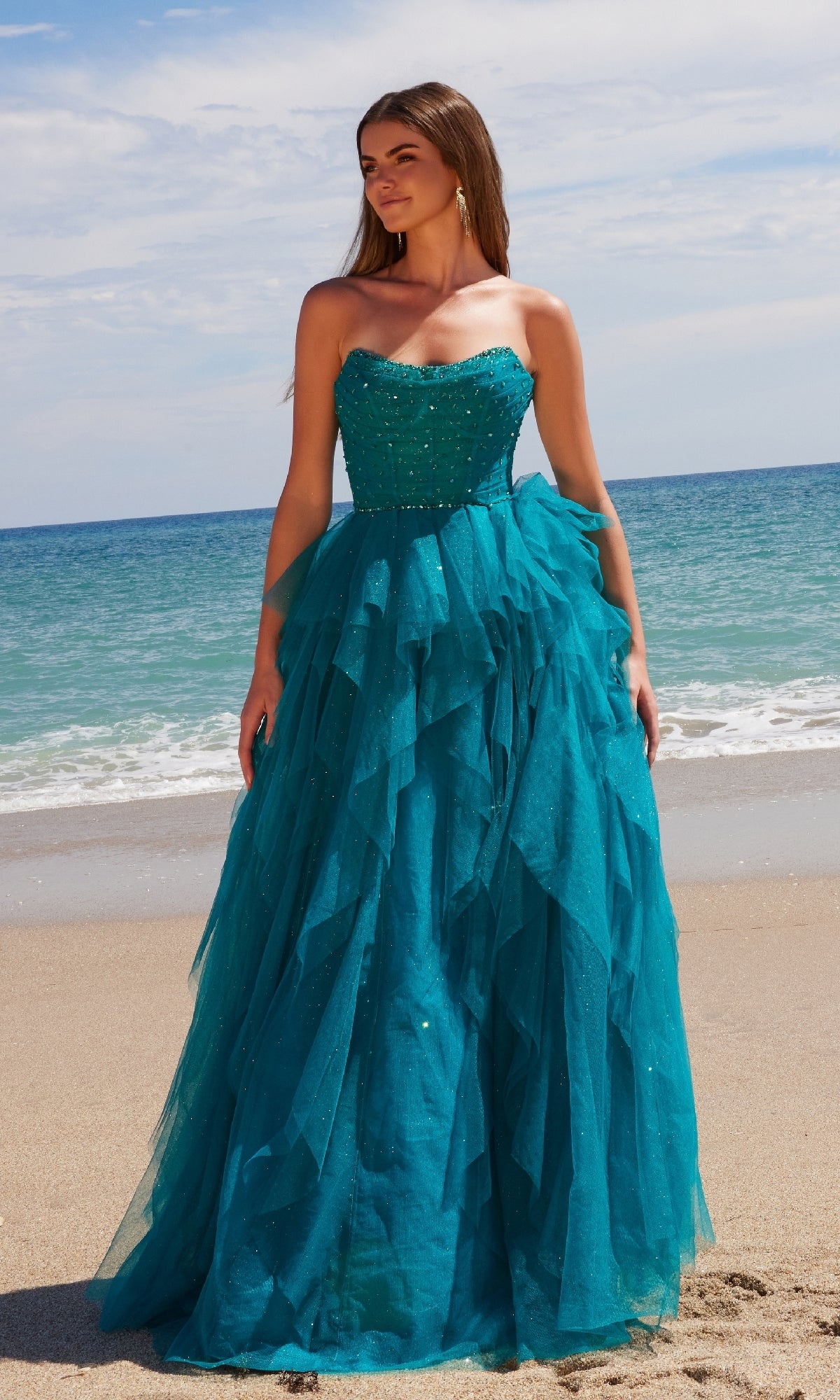 A woman in the Nox Anabel A1591 Long Prom Dress, a strapless corset gown with a ruffled A-line skirt, stands on a sandy beach with teal fabric flowing against ocean waves and a blue sky.