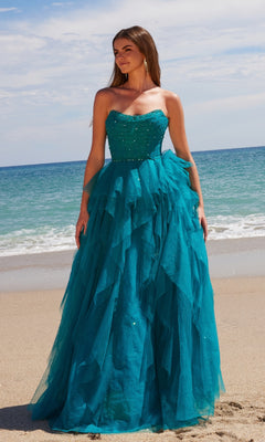 A woman in the Nox Anabel A1591 Long Prom Dress, a strapless corset gown with a ruffled A-line skirt, stands on a sandy beach with teal fabric flowing against ocean waves and a blue sky.
