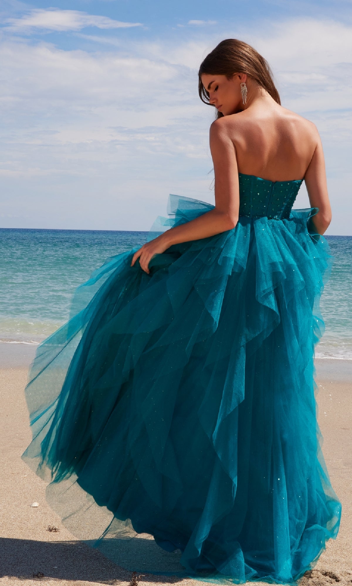 Wearing the Nox Anabel A1591 long prom dress with a glitter tulle ruffled A-line skirt, a woman stands on a sandy beach, gazing at the ocean beneath a partly cloudy sky.