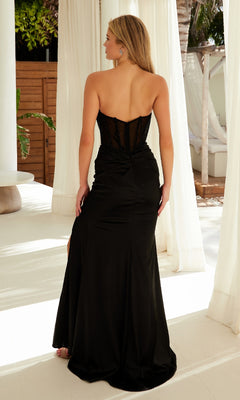 A woman in the Nox Anabel A1596 Strapless Long Glitter Prom Gown with a sheer back stands indoors, facing away, surrounded by white curtains and natural light.