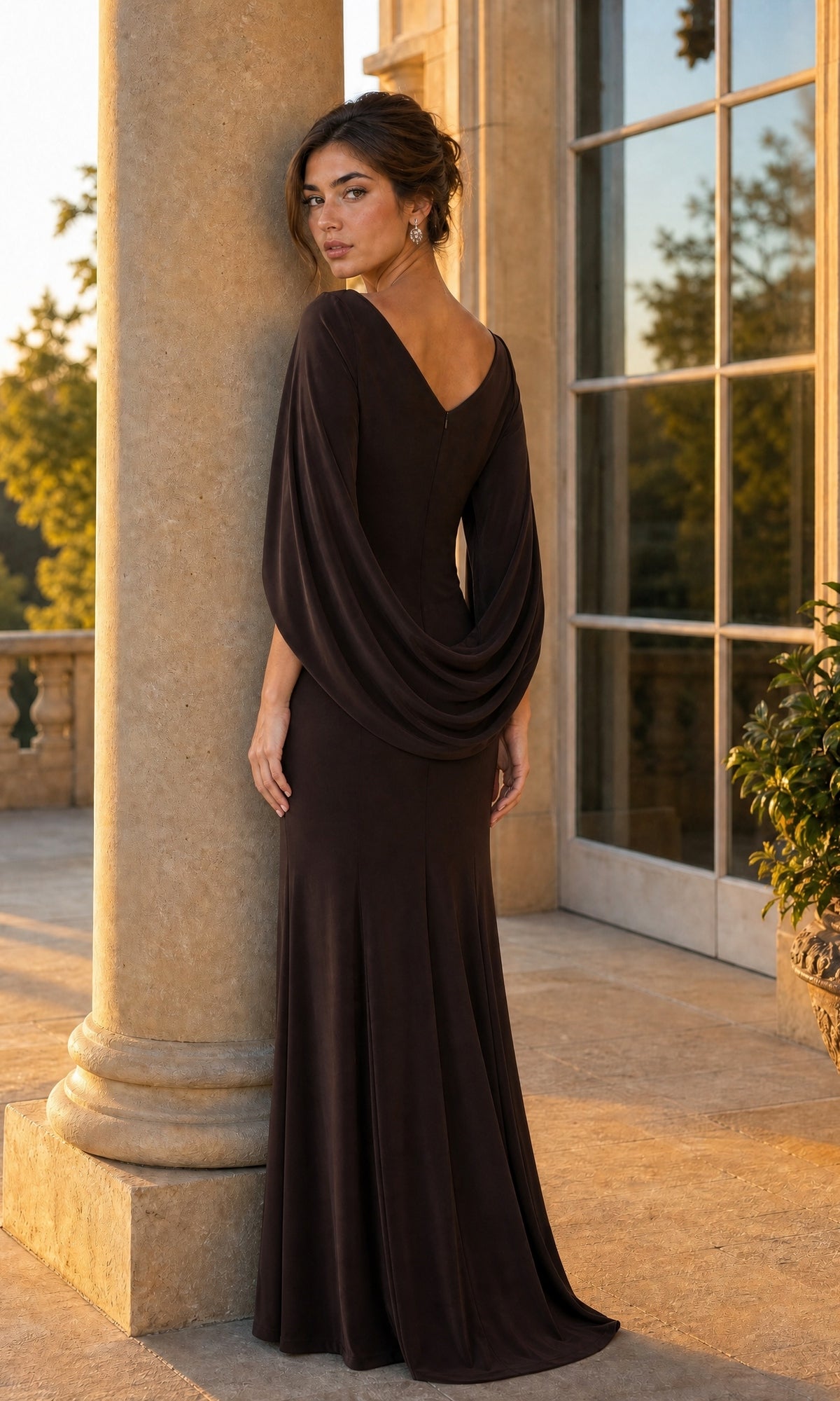 A woman in a long brown Betsy & Adam A23033 formal dress with a draped back stands near a column on a patio, looking over her shoulder.