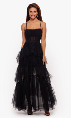 A woman stands smiling in the Betsy and Adam Long Ruffled Prom Dress A24201—a sleeveless black tulle gown with thin straps, sheer bodice, tiered skirt—and black open-toe heels, set against a plain white background.