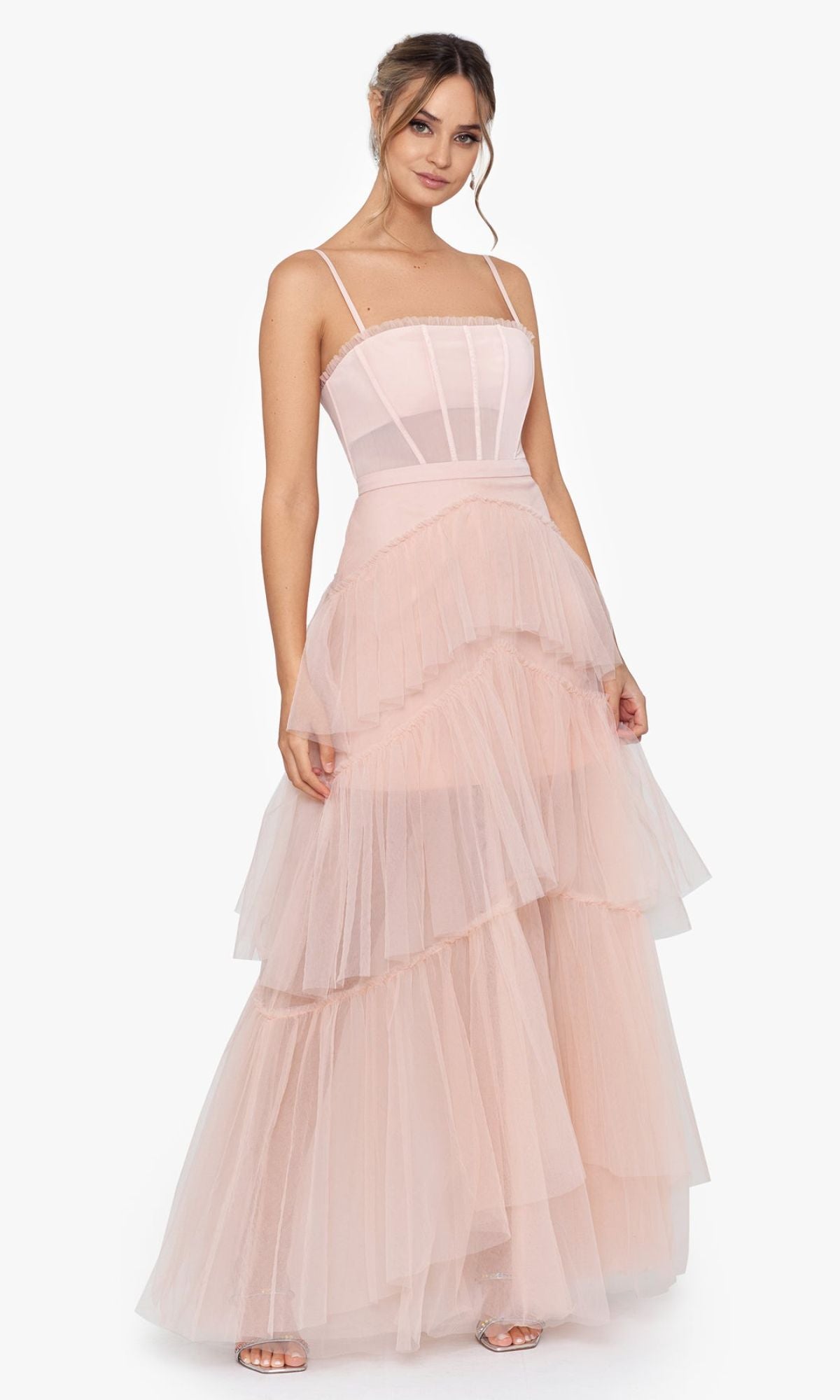A woman wears the Betsy and Adam Long Ruffled Prom Dress A24201 in light pink, featuring spaghetti straps, a fitted bodice, and tiered tulle layers, posed against a plain white background.