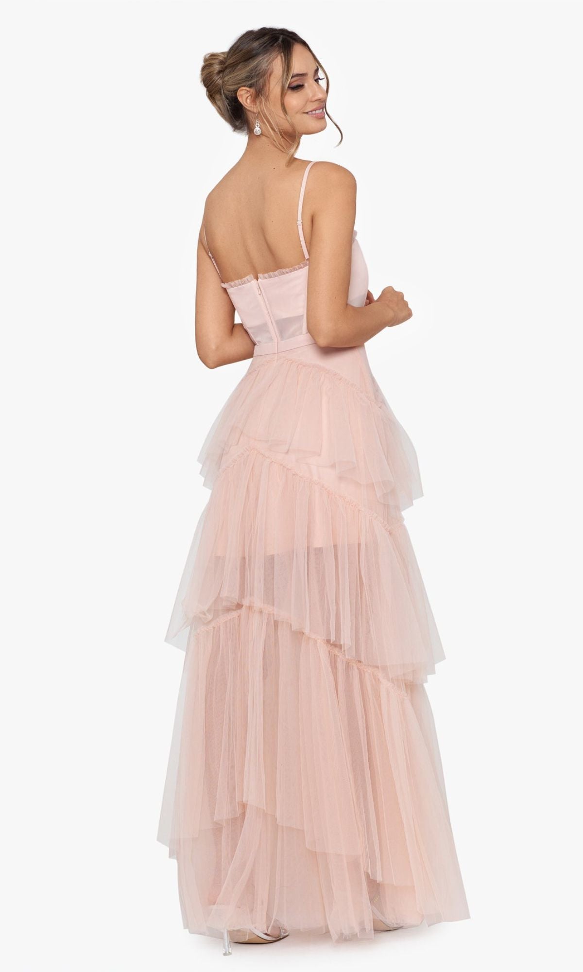 A woman stands smiling over her shoulder in the Betsy and Adam Long Ruffled Prom Dress A24201, a light pink tiered formal dress with thin straps, set against a plain white background.