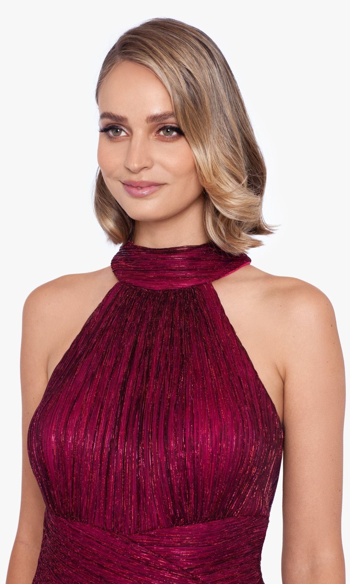 A woman with short, wavy blonde hair wears the Empire-Waist High-Neck Long Shimmer Prom Dress A24948 in pleated burgundy, featuring a sleeveless design and subtle shimmer, standing against a plain light background.