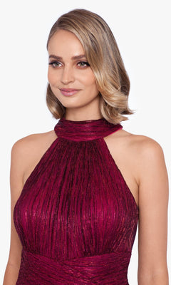 A woman with short, wavy blonde hair wears the Empire-Waist High-Neck Long Shimmer Prom Dress A24948 in pleated burgundy, featuring a sleeveless design and subtle shimmer, standing against a plain light background.
