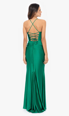 A woman stands facing away, wearing the Betsy and Adam Long Green Prom Dress A25610—a long emerald satin gown with a cowl neck and crisscross lace-up back detailing.