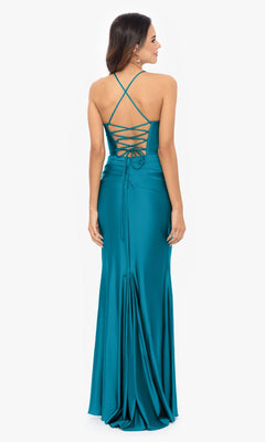 A woman stands facing away from the camera in the Betsy and Adam Long Green Prom Dress A25610, featuring a corset-style bodice, crisscross lace-up back, and a floor-length teal hem.