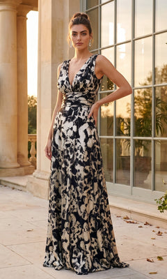 A woman in a sleeveless, floor-length navy and gold floral Betsy & Adam A26172 formal dress stands outside a building with tall columns and large windows at sunset.
