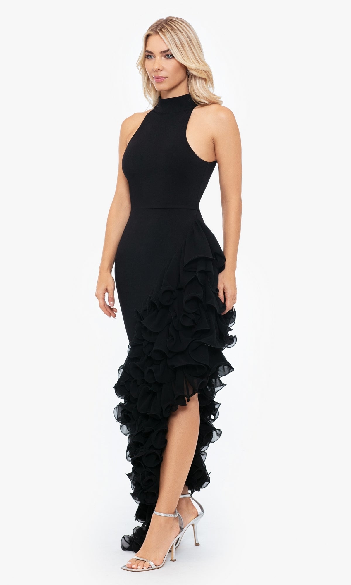 A woman wears the Betsy and Adam Long Black Ruffled Prom Dress A26173, featuring a high neckline, sleeveless bodice, and asymmetrical ruffled skirt, paired with silver high-heeled sandals.