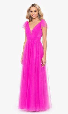 A woman stands and smiles against a white background, wearing the Betsy & Adam Shoulder-Bow Long Prom Dress A26256— a floor-length bright pink gown with cap sleeves, a deep V-neckline, and a fitted waist.