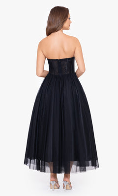 A woman stands in high-heeled sandals, facing away to show the fitted corset bodice and visible back zipper of the Black Strapless Tea Length Wedding-Guest Dress—an ideal chic choice for any wedding guest.