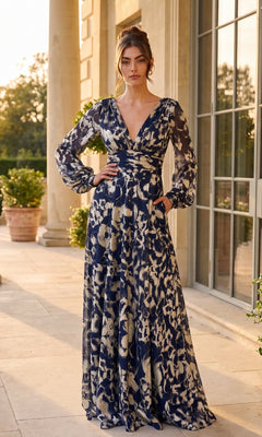 A woman stands outdoors next to large windows and columns, wearing the Betsy & Adam A26583 long formal dress. This navy gown features a gold metallic foil print, long sleeves, and a deep V-neck. Its elegant A-line silhouette enhances the graceful appeal of the stunning floral design.