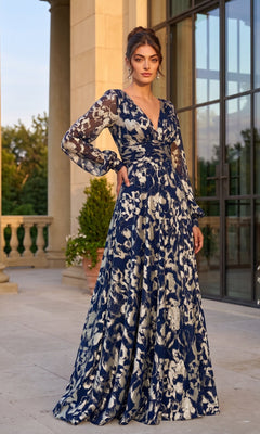 A woman wearing the Betsy & Adam A26583 long formal dress in Navy/Gold, featuring a floral design with sheer sleeves and an elegant A-line silhouette, stands outside a building with columns and large windows.