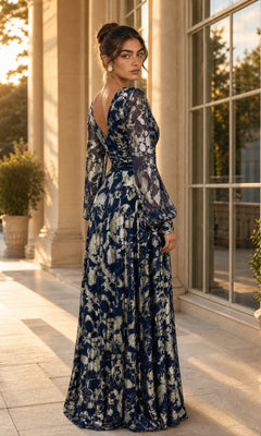 A woman stands on a sunlit terrace, looking over her shoulder, wearing the Betsy & Adam A26583 Long Formal Dress—a navy gown with an elegant A-line silhouette, gold metallic foil print, and sheer sleeves.