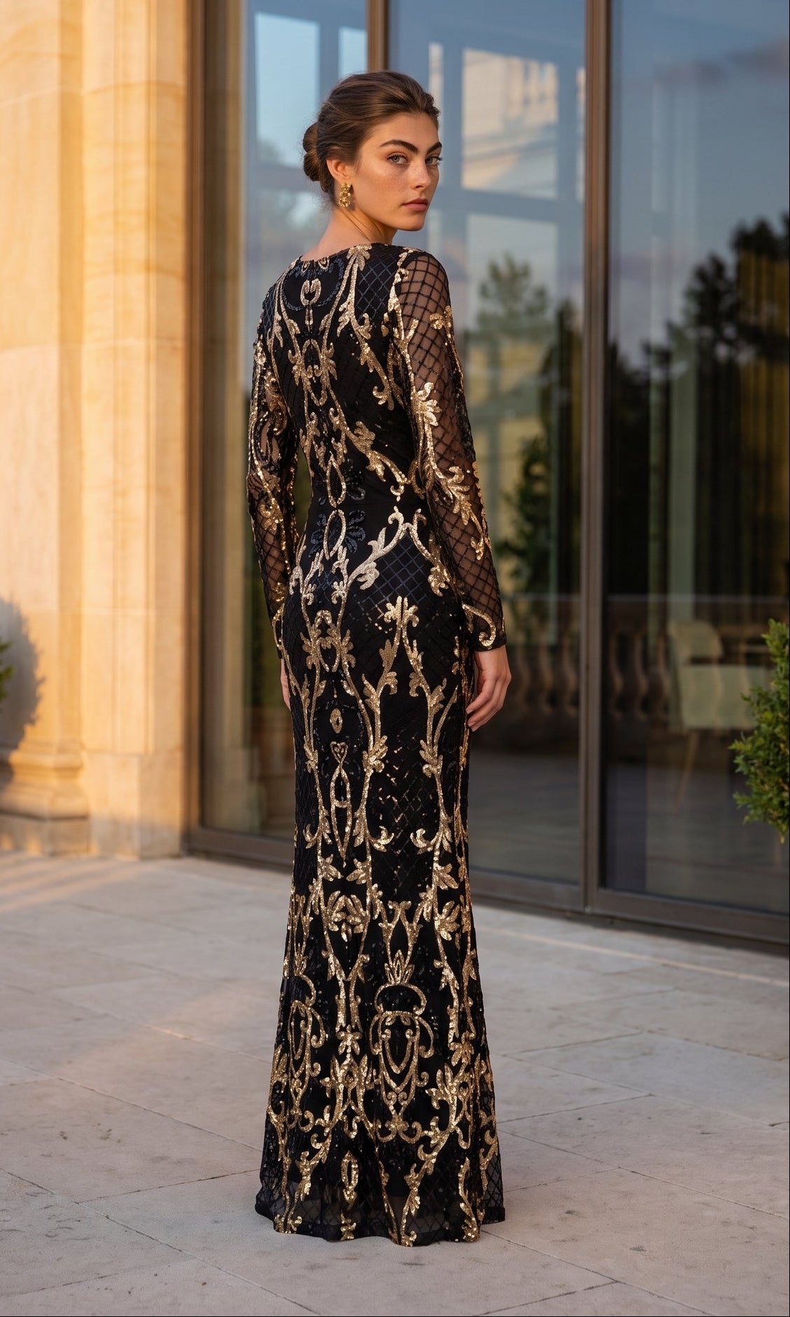 A person stands outdoors in front of a glass building, looking back over their shoulder while wearing the Betsy & Adam A26776 long formal dress, which features black fabric with gold ornate patterns and sheer sleeves.