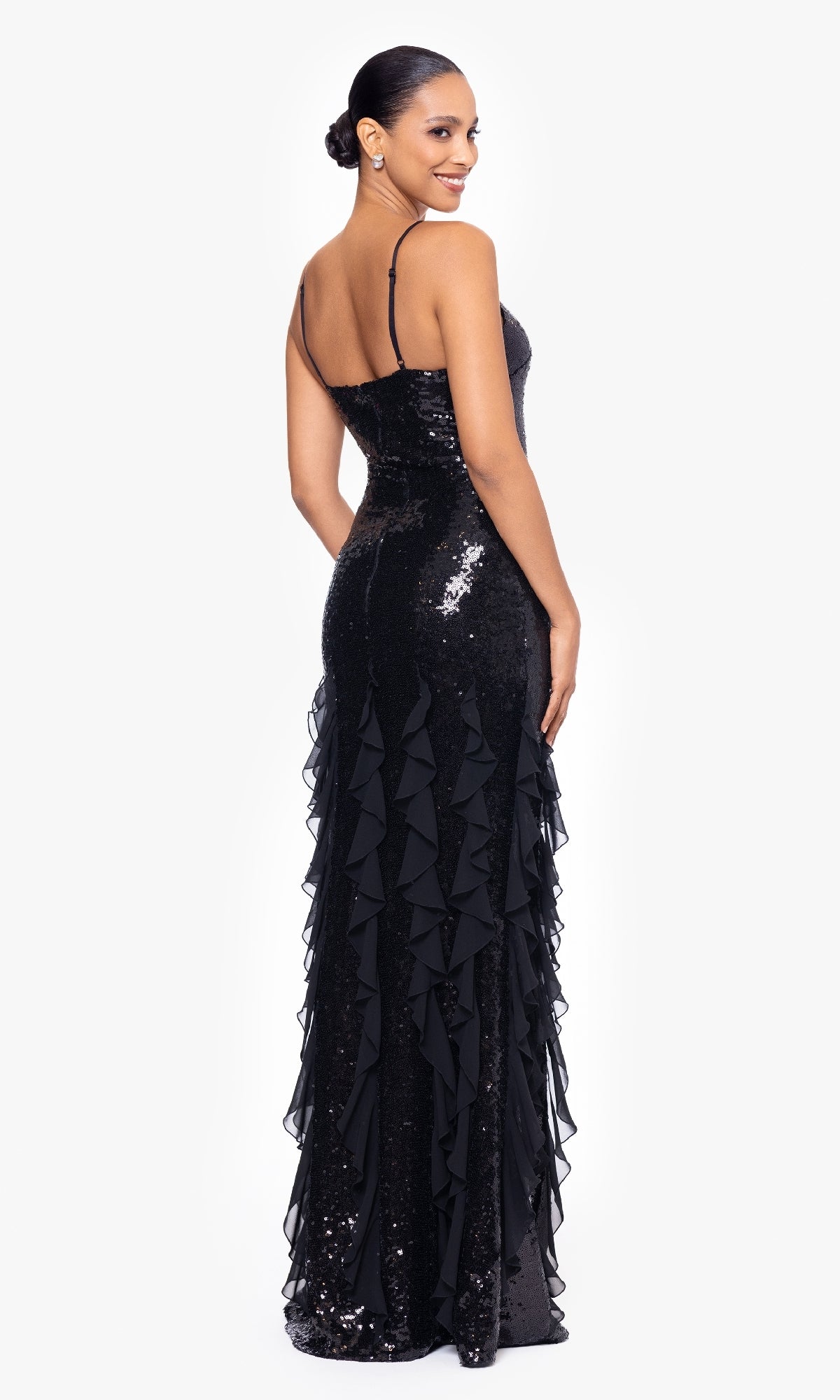 A woman wears the Betsy & Adam A26805 Long Black Sequin Formal Gown, featuring thin straps and ruffle details, as she stands facing backward and smiles over her shoulder.