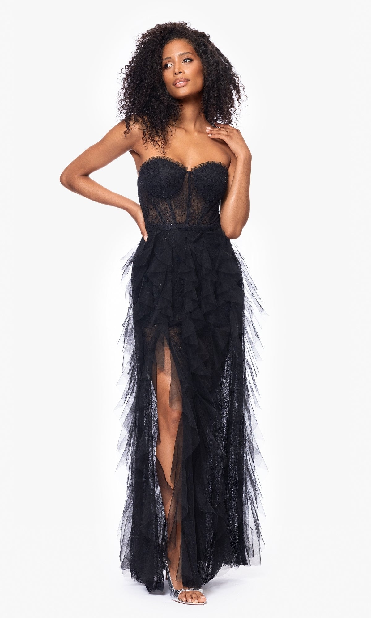 Woman wearing the Betsy & Adam A26845 Strapless Black Prom Gown featuring lace, sheer ruffled tulle layers, and a thigh-high slit, standing against a plain white background.