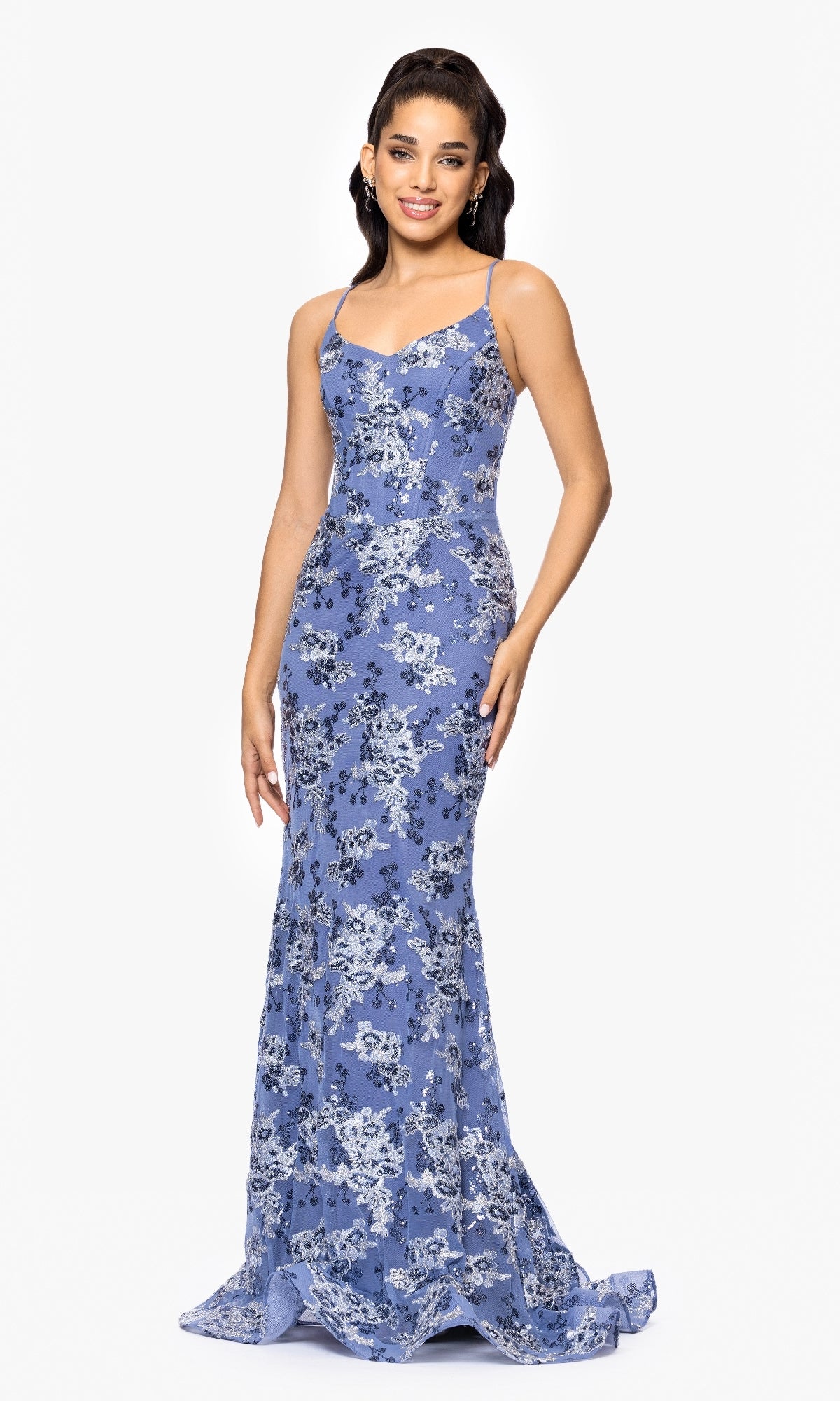 A woman smiles at the camera against a white background, wearing the Betsy and Adam A26917, a long fitted formal dress in light blue floral lace with thin straps and a lace-up back. She has long dark hair.