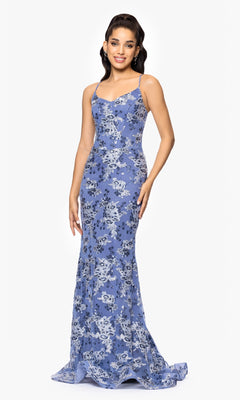 A woman smiles at the camera against a white background, wearing the Betsy and Adam A26917, a long fitted formal dress in light blue floral lace with thin straps and a lace-up back. She has long dark hair.