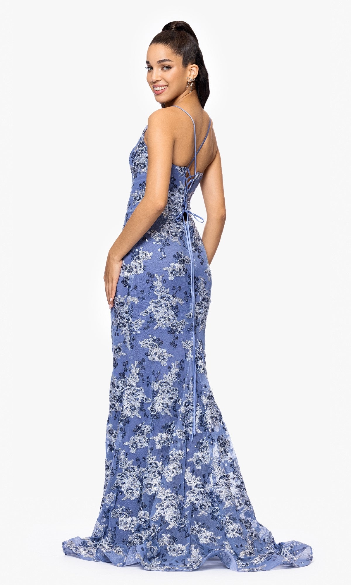 A woman models the Betsy and Adam A26917 long formal dress, featuring sleeveless light blue floral lace and a lace-up back, smiling over her shoulder against a white background.
