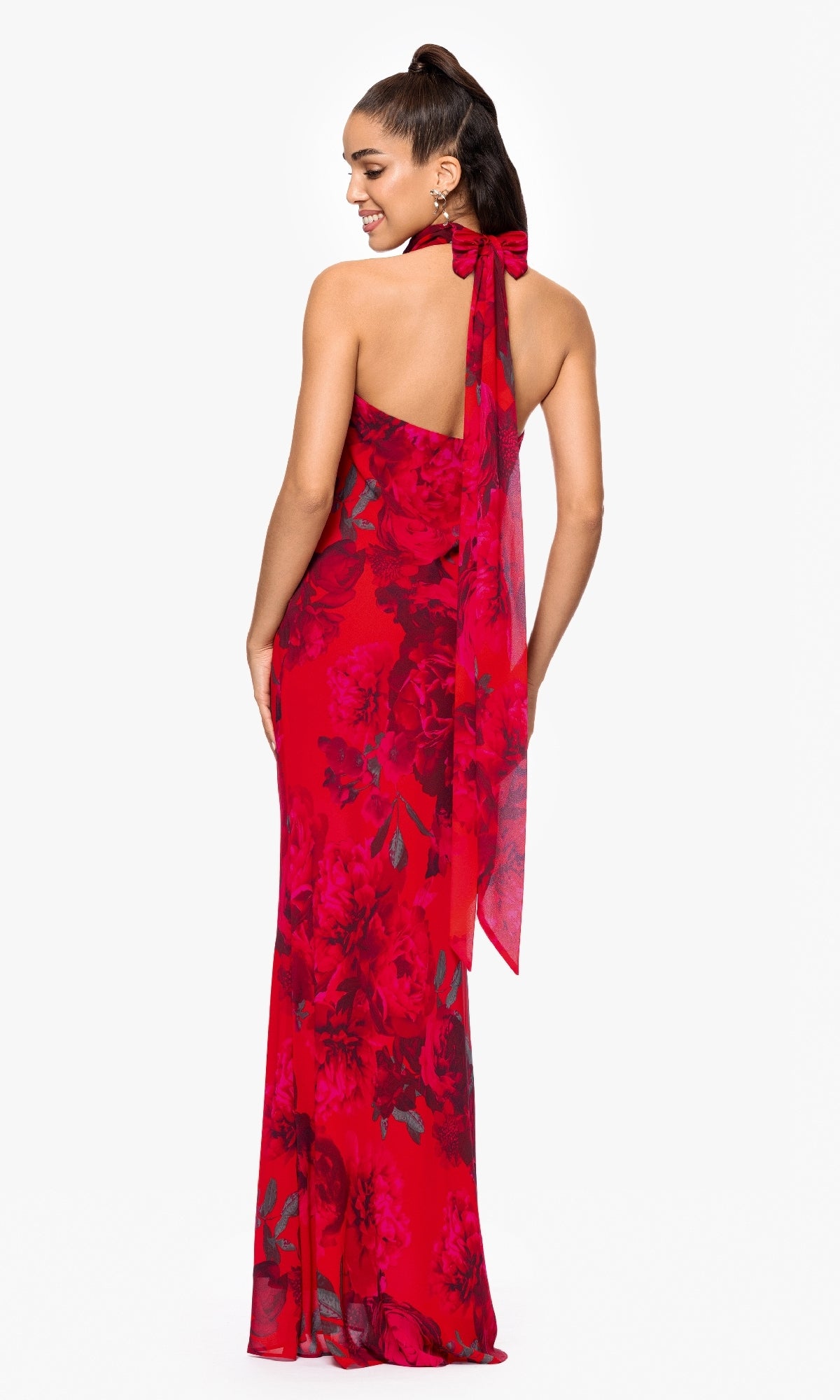 A woman with a ponytail faces away, wearing the Betsy & Adam A26926 long red floral formal dress featuring a halter neckline and a large bow at the neck.