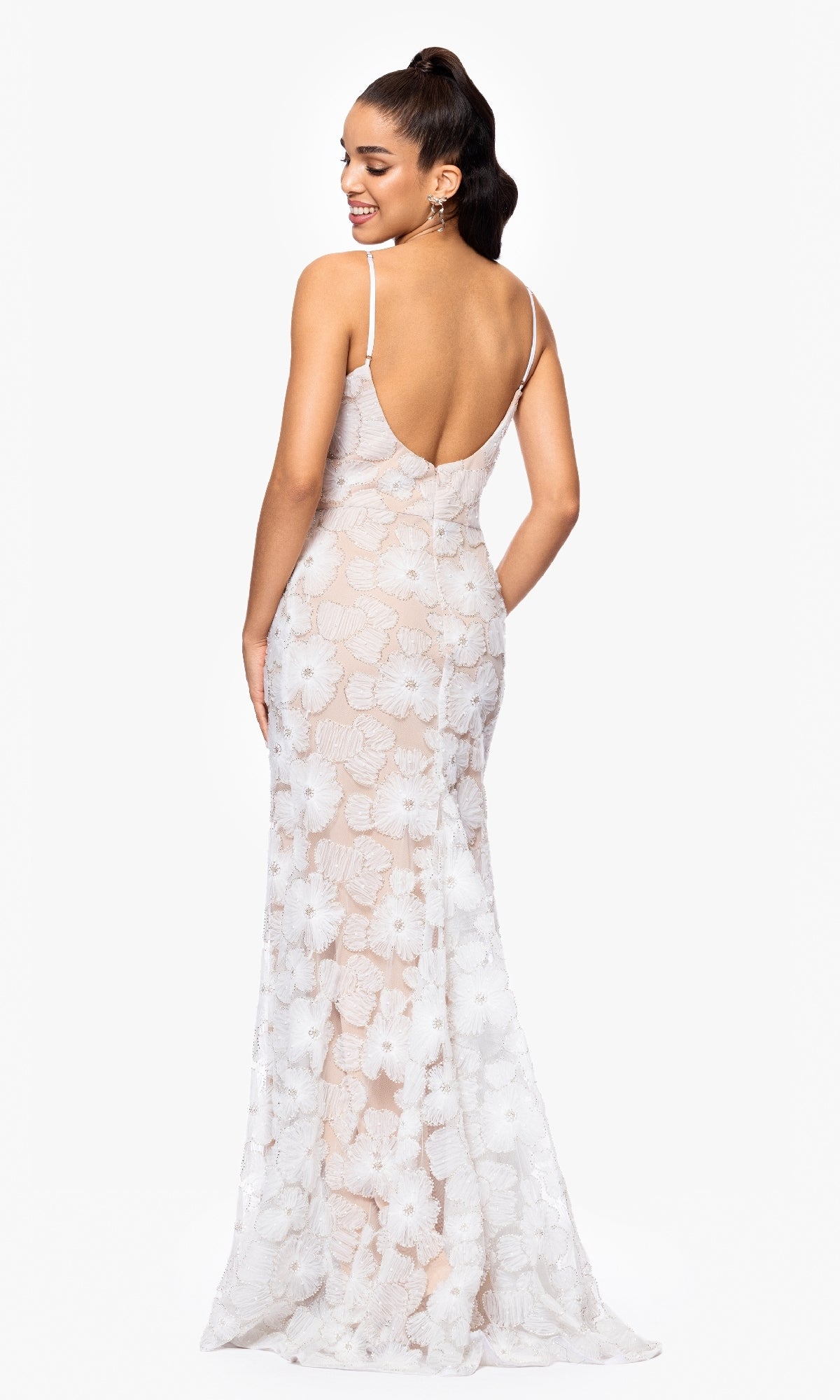 Woman in the Betsy and Adam A26948 long formal dress, fitted white with floral pattern, delicate lace, and adjustable straps, faces away and looks over her shoulder against a plain white background.