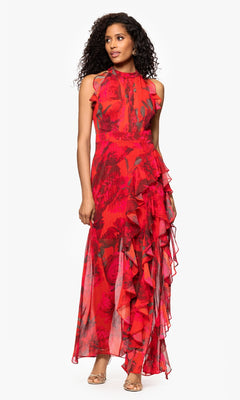 A woman stands wearing the Betsy and Adam A26962 Floral-Print Red Formal Dress, a sleeveless, floor-length gown with ruffle details, paired with open-toed heels.