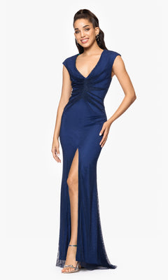A woman models the Betsy and Adam A26963 long formal dress—sleeveless, navy blue with a v-neckline and thigh-high slit—paired with silver heels, standing against a white background.