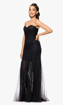 A woman stands and smiles, posing in the Betsy and Adam A26972 long formal dress— a strapless black mermaid gown with a sweetheart neckline, illusion skirt, and subtle sparkly details—against a plain white background.