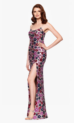 A woman in the Betsy & Adam A27017 Floral-Sequin Long Prom Dress, featuring a sleeveless design, colorful patterns, and a thigh-high slit, stands smiling against a plain background.