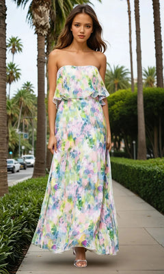 A woman in a Betsy & Adam A27018 Strapless Wedding Guest Gown walks on a sunlit, palm-lined sidewalk.