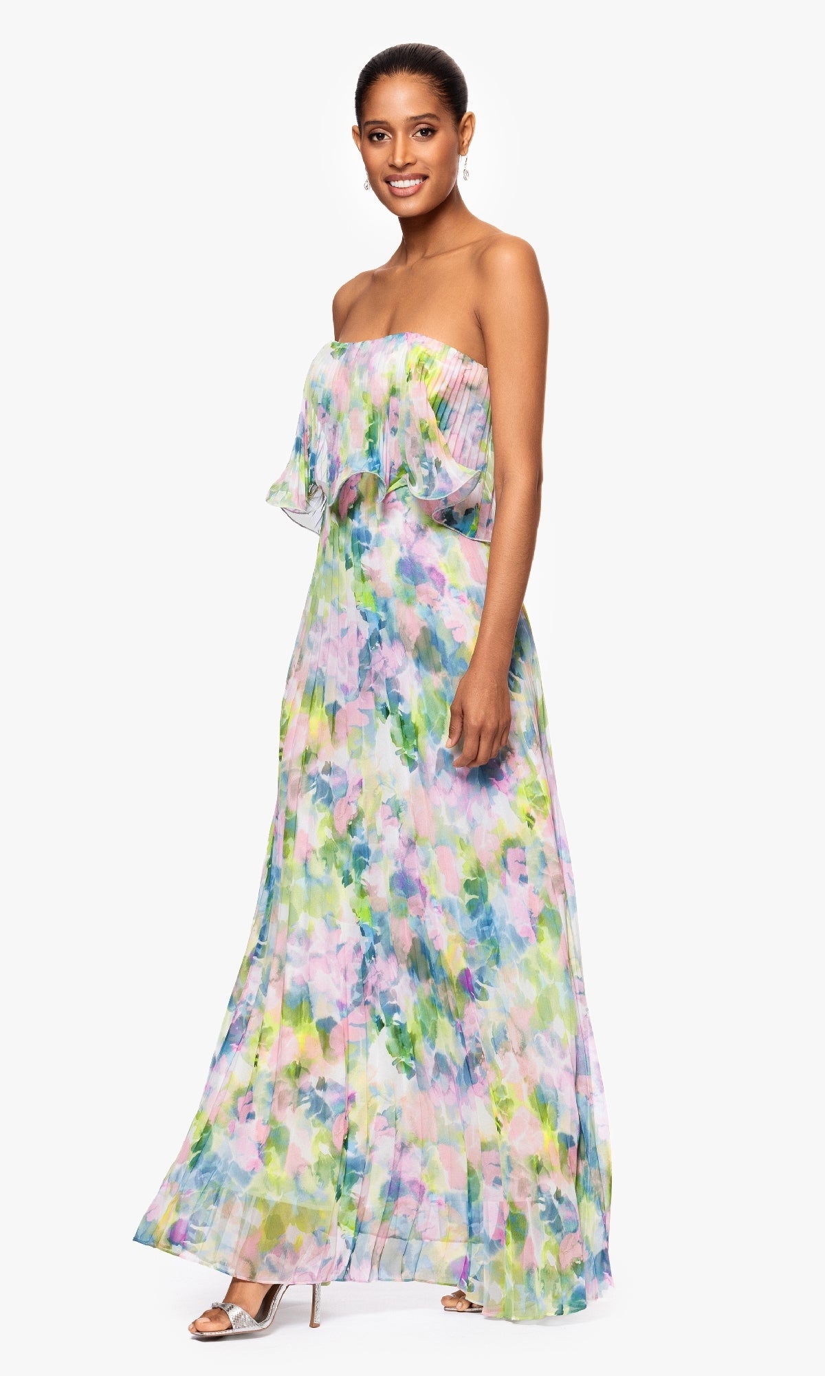 A woman wears the Betsy & Adam A27018 Strapless Wedding Guest Gown with pastel watercolor florals and silver heels, standing against a white background—an elegant option for wedding guests.