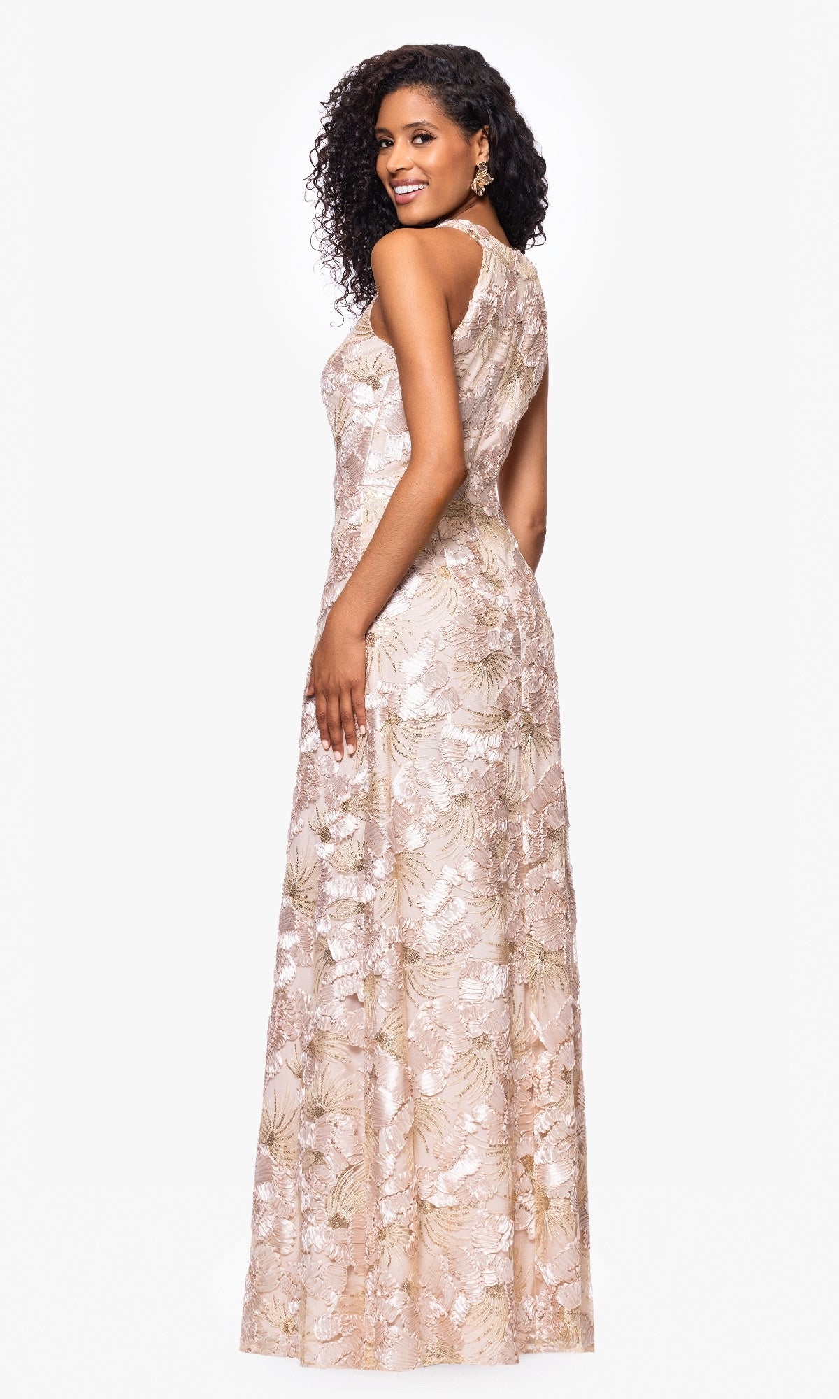A woman stands against a plain white background, looking over her shoulder in the Betsy and Adam A27023 long formal dress—a sleeveless, floor-length gown featuring light-colored floral soutache lace.