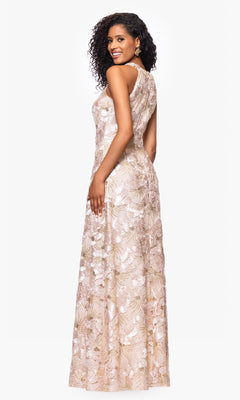 A woman stands against a plain white background, looking over her shoulder in the Betsy and Adam A27023 long formal dress—a sleeveless, floor-length gown featuring light-colored floral soutache lace.