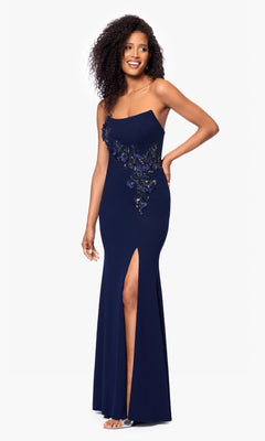 A woman stands smiling against a white background, wearing the Long Formal Dress: Betsy and Adam A27030, a navy blue, form-fitting gown with a beaded bodice, floral sequin details, and a thigh-high slit.