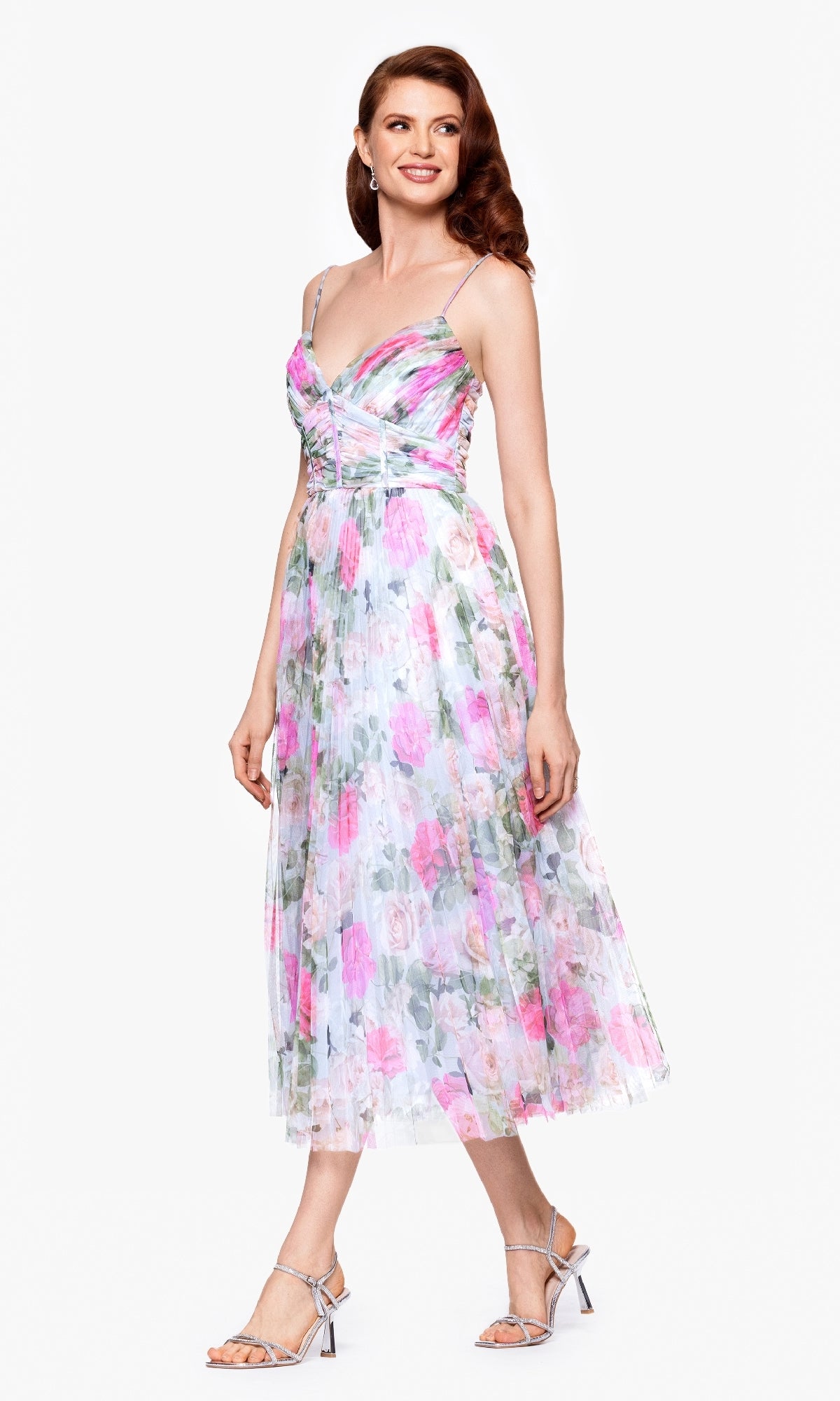 A woman stands smiling against a plain background in the Betsy & Adam A27054 Print Wedding Guest Dress, featuring a floral tea-length design with a fitted bodice, flared skirt, and strappy heels.