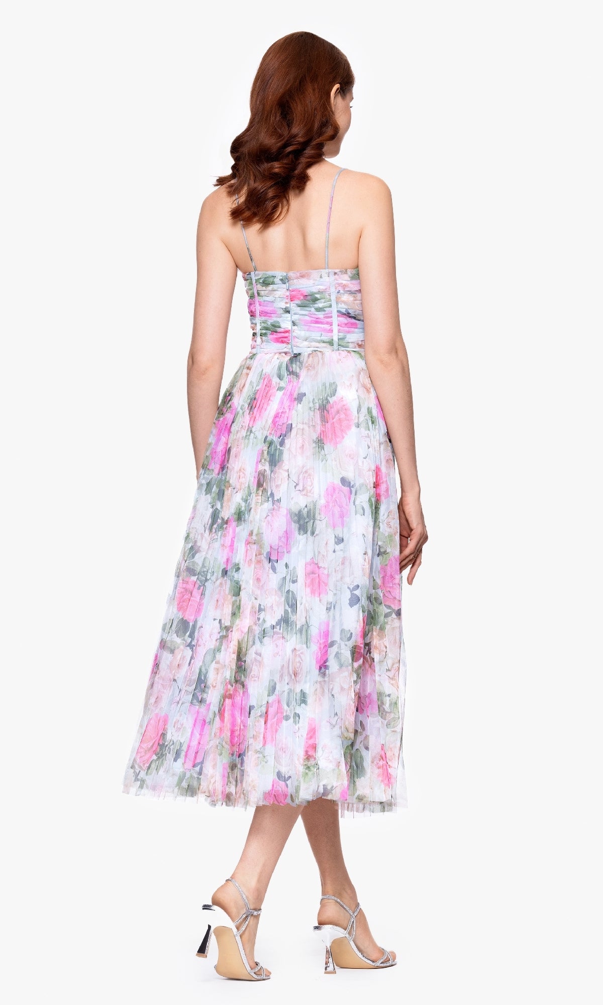 A woman stands facing away, wearing the Betsy & Adam A27054 Print Wedding Guest Dress — a pink and green floral tea-length dress with thin straps and pleated skirt — paired with strappy high-heeled sandals.