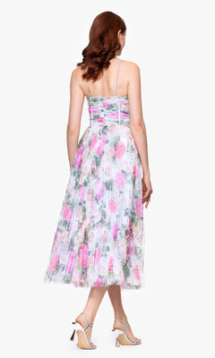 A woman stands facing away, wearing the Betsy & Adam A27054 Print Wedding Guest Dress — a pink and green floral tea-length dress with thin straps and pleated skirt — paired with strappy high-heeled sandals.