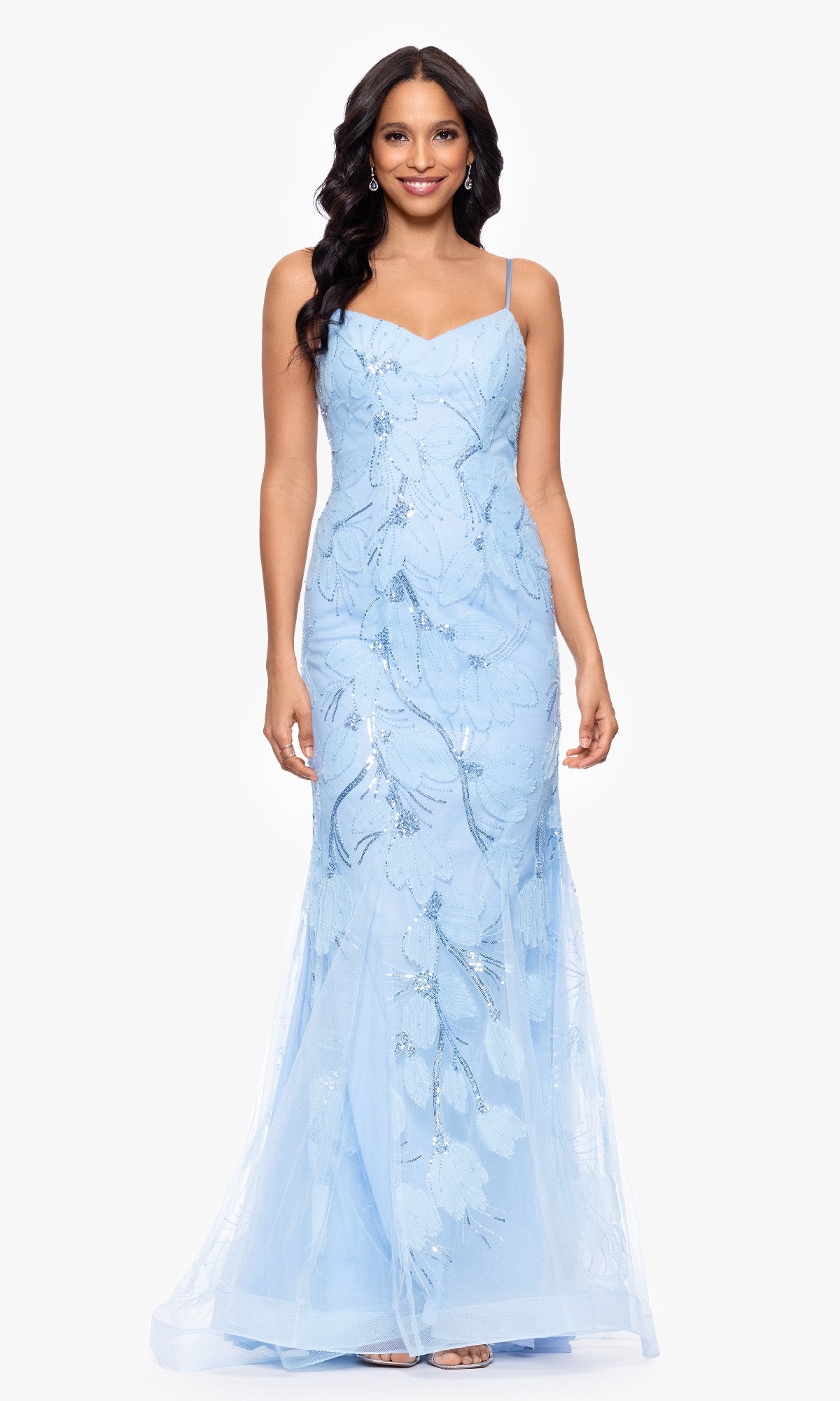 A woman stands facing forward against a white background, wearing the Betsy and Adam A27097 long formal dress in light blue with thin straps and floral sequin embellishments.