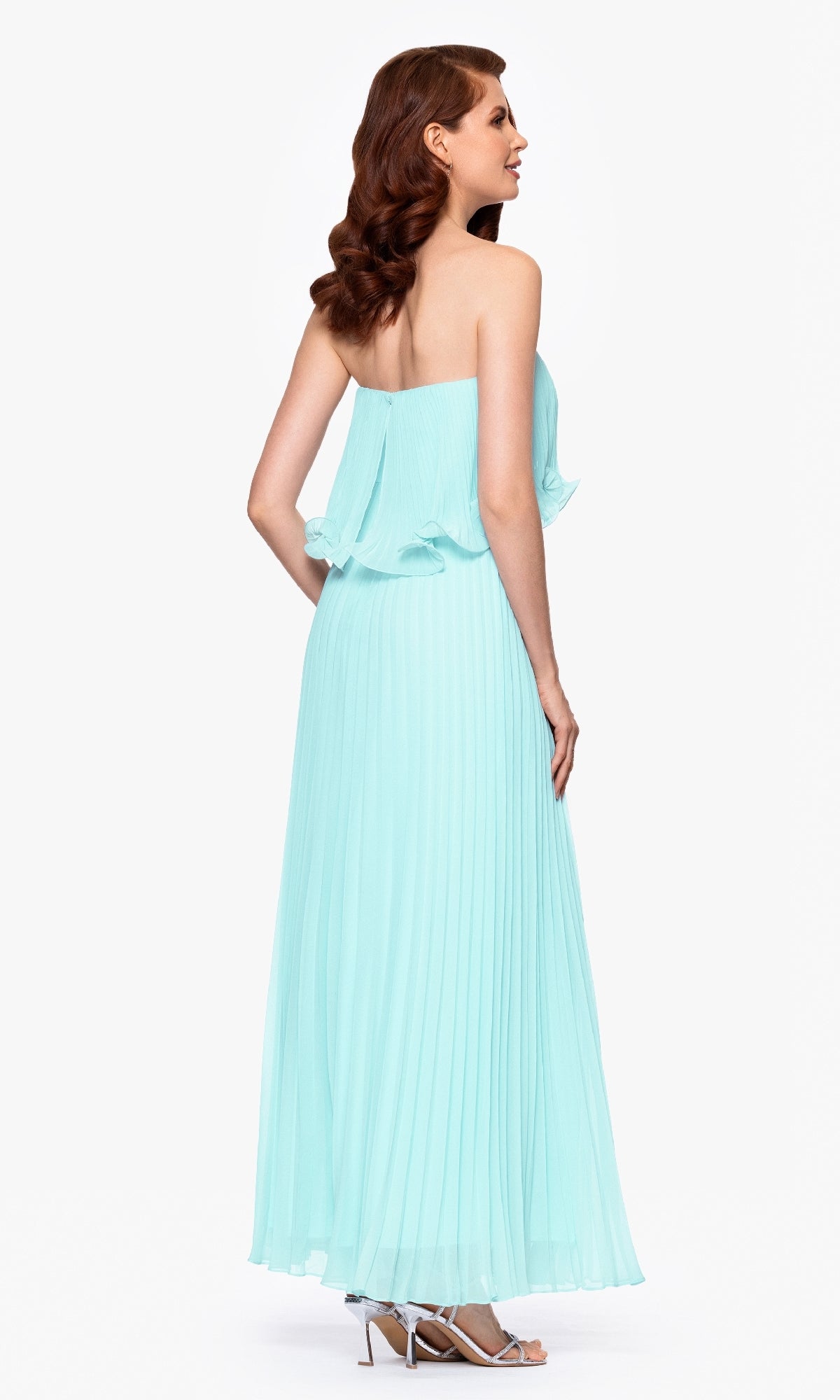 A woman in profile wears the Betsy & Adam A27098 Strapless Wedding Guest Dress—floor-length, light blue, and pleated—with silver heels, standing against a white background.