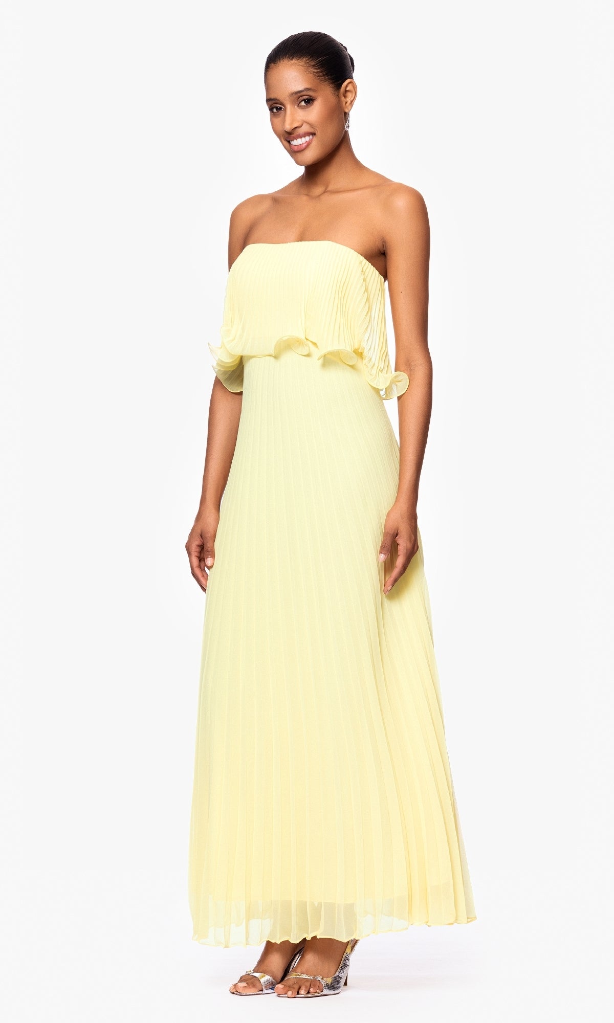 A woman stands against a plain white background wearing the Betsy & Adam A27098 strapless, floor-length, pleated light yellow wedding guest dress with clear high-heeled sandals.