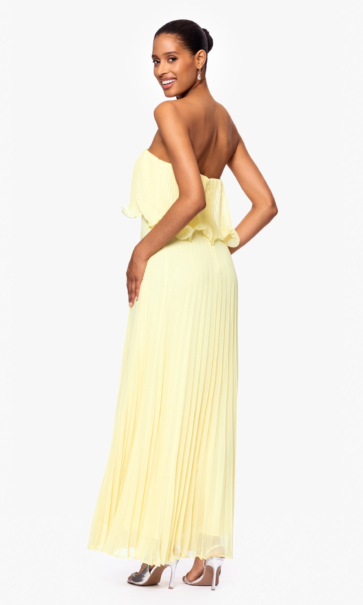 A woman in the Betsy & Adam A27098 strapless wedding guest dress, light yellow with pleats, and silver heels stands facing away, looking over her shoulder and smiling.