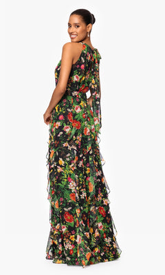 A woman smiles while standing slightly sideways in the Betsy & Adam A27102 Floral Wedding Guest Dress—a sleeveless, floor-length black gown with floral print, ruffles, and a pleated bodice—against a white background.