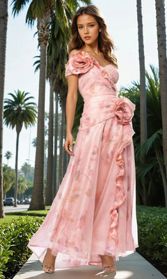 A woman stands outdoors among palm trees, wearing the Betsy & Adam A27103 long formal dress, featuring a pink floral print and large rosette details on the shoulder and waist.